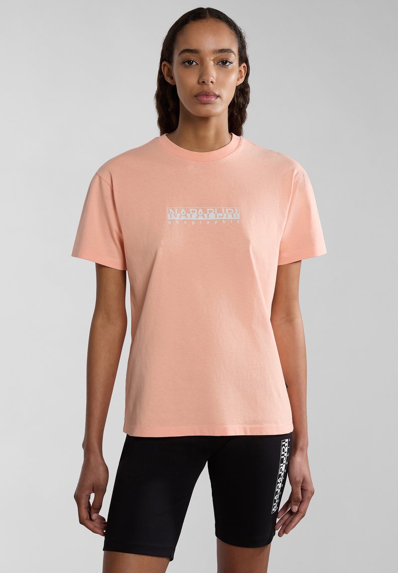 Hellrosa Baumwoll-T-Shirt mit kurzen Ärmeln, mit einem weißen gedruckten Logo auf der Vorderseite. Kombiniert mit schwarzen, eng anliegenden Shorts.