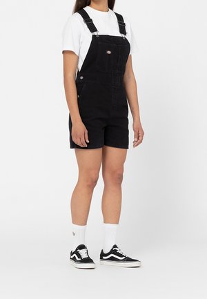 Zwarte denim tuinbroeken met een relaxte pasvorm, voorzien van verstelbare schouderbanden, een voorzak en zijknoppen. Gecombineerd met een witte shirt en zwarte sneakers.