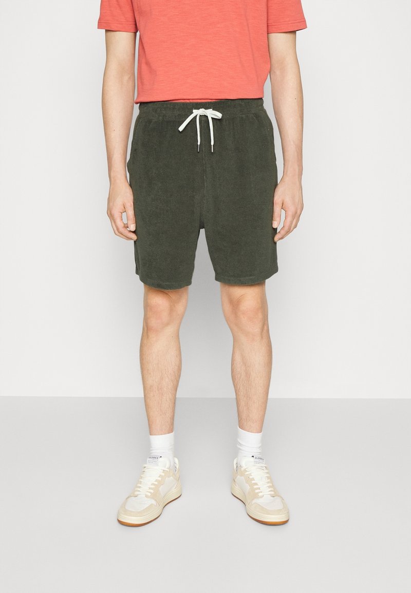 Shorts verts texturés avec une taille élastique et un cordon blanc, associés à un t-shirt corail et des baskets claires.