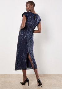 Granatowa sukienka wyszywana cekinami z krótkimi rękawkami, rozcięciem do uda i otworem w kształcie dziury na plecach. Noszona z czarnymi szpilkami.