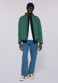 Zelená péřová bunda s vysokým límcem, spárovaná s tmavým tričkem a bílým tričkem s potiskem. Modré rovné džíny a zelené tenisky.