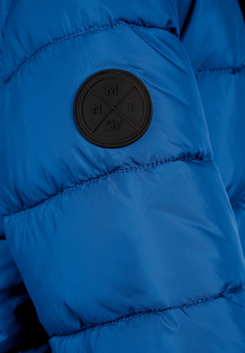 Veste matelassée bleue avec une texture brillante. Dotée d'un patch noir rond avec du texte en relief, situé sur la partie supérieure de la manche.