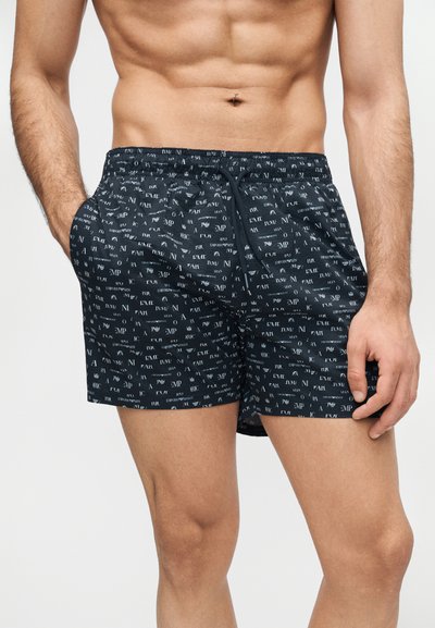 Άνδρας που φοράει μαύρα μαγιό με λευκό τυπωμένο κείμενο διάσπαρτο, ελαστική μέση με κορδόνι, στέκεται με τα χέρια κοντά στις τσέπες.