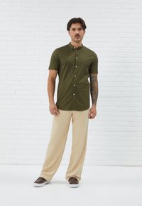 Olivenfarvet kortærmet skjorte med button-down krave, der har en brystlomme. Sammen med beige bredbenede bukser og brune sko.