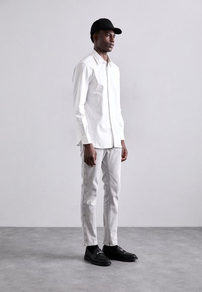 Chemise blanche à boutons, jean slim gris, mocassins noirs et casquette noire. Le modèle se tient contre un fond gris clair.