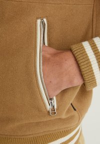 Brązowa polarowa kurtka ma zamek błyskawiczny w bocznej kieszeni z kremowym suwakem. Rękawy mają prążkowaną strukturę w brązowo-kremowe paski.