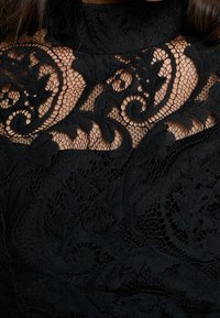 Le tissu en dentelle noire présente des motifs floraux et tourbillonnants complexes, avec une surface texturée. Le col haut met en valeur le design détaillé.