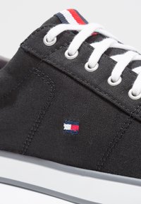 Sneaker nera in tela con lacci bianchi, asole in metallo, striscia rossa e bianca sulla linguetta e piccolo logo ricamato in rosso, bianco e blu.