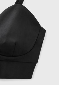 Bonnet de soutien-gorge rembourré en tissu noir avec une large bande sous la poitrine et une bretelle étroite réglable sur un fond clair.