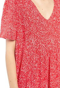 Robe rouge avec un motif à pois blancs, à manches courtes et décolleté en V portée par une personne montrant ses bras et une partie de la poitrine.