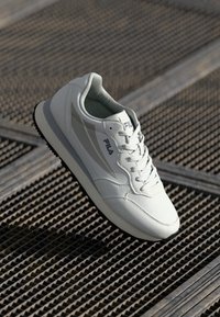 White sneaker with a smooth synthetic upper, rounded toe, and minimalistic design. Features logo and accents in light gray, textured sole.