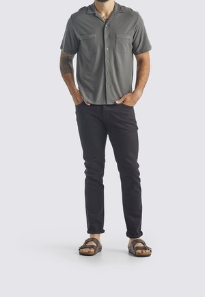 Man draagt een grijs overhemd met korte mouwen en knopen, zwarte opgerolde broek, bruine sandalen, staat met handen in de zakken tegen een effen achtergrond.