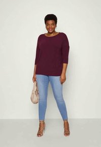 Tumeroheline kolmeveerandvarrukaga särk, kombineeritud hele siniste kitsaste teksadega ja hõbedate rihmlike kontsadega, hoides käes beeži käekotti. Sile tekstuur.