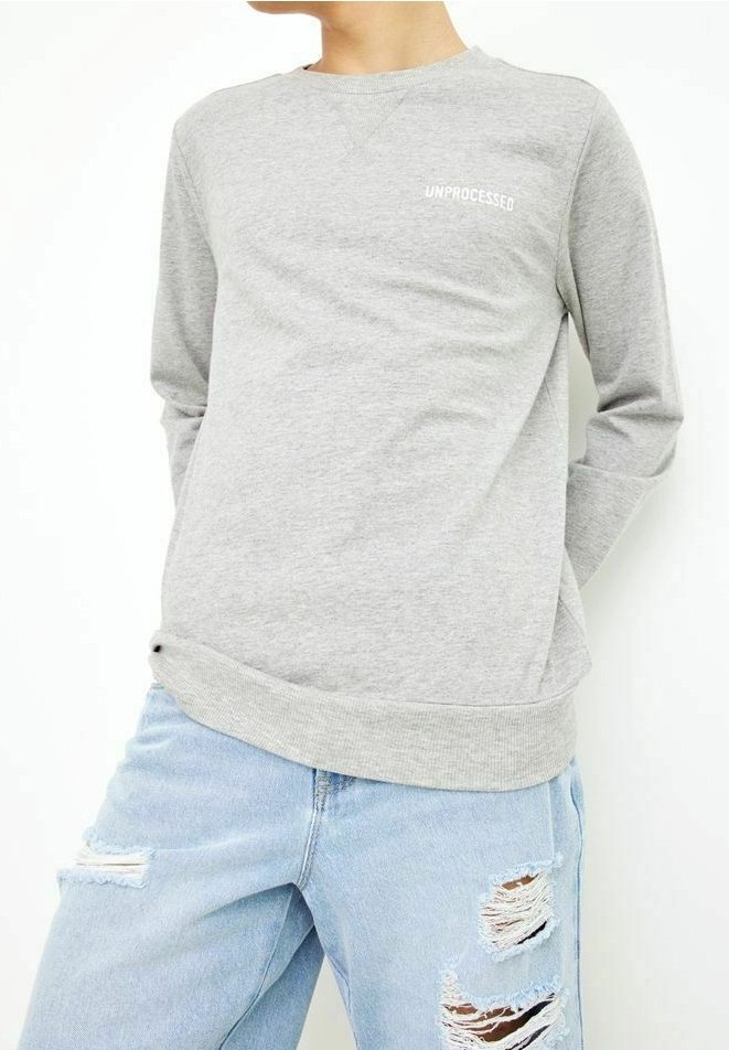Personne portant un sweat-shirt gris clair avec le texte "UNPROCESSED" et un jean déchiré bleu clair, debout les mains derrière le dos.