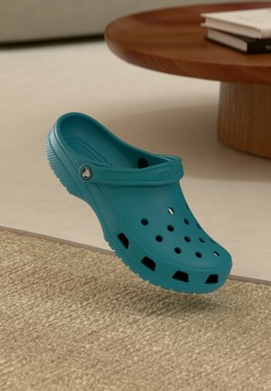 Blauer leichter Clogs mit Belüftungslöchern und verstellbarem Fersenriemen, liegen auf einem beigen gewebten Teppich neben einem Holztisch mit Büchern.