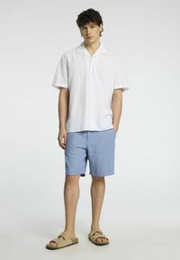 Bílá košile s krátkým rukávem a knoflíkovou légou, spárovaná se světle modrými šortkami a hnědými sandály. Hladká tkanina, volný střih, ležérní design.