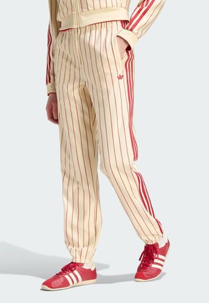 Persoon draagt crèmekleurige trainingsbroek met rode verticale strepen, rode zijstrepen en rode sneakers met witte strepen.