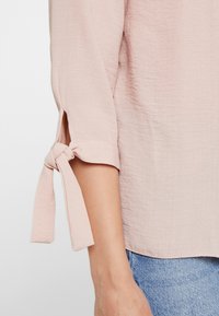 Blusa rosa chiaro con finitura testurizzata. Presenta maniche arrotolate con lacci ai polsi e una vestibilità ampia. Abbinata a jeans blu.