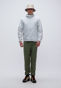 Άνδρας που φορά ανοιχτό γκρι μπουφάν με κουκούλα, χακί παντελόνι, μπεζ καπέλο τύπου bucket και μαύρα και κίτρινα παπούτσια για πεζοπορία, στέκεται μπροστά σε απλό φόντο.