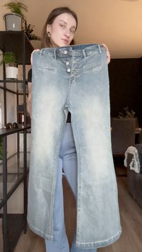 Jeans de denim azul claro con un diseño ancho y dividido. Presenta cinco botones y bolsillos frontales. Textura descolorida en las piernas con acentos cosidos.