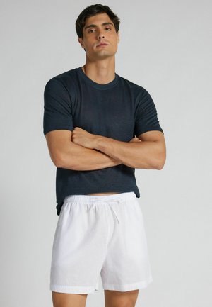 Uomo che indossa una t-shirt blu scuro con pantaloncini bianchi con coulisse, in piedi con le braccia incrociate davanti a uno sfondo chiaro e uniforme.