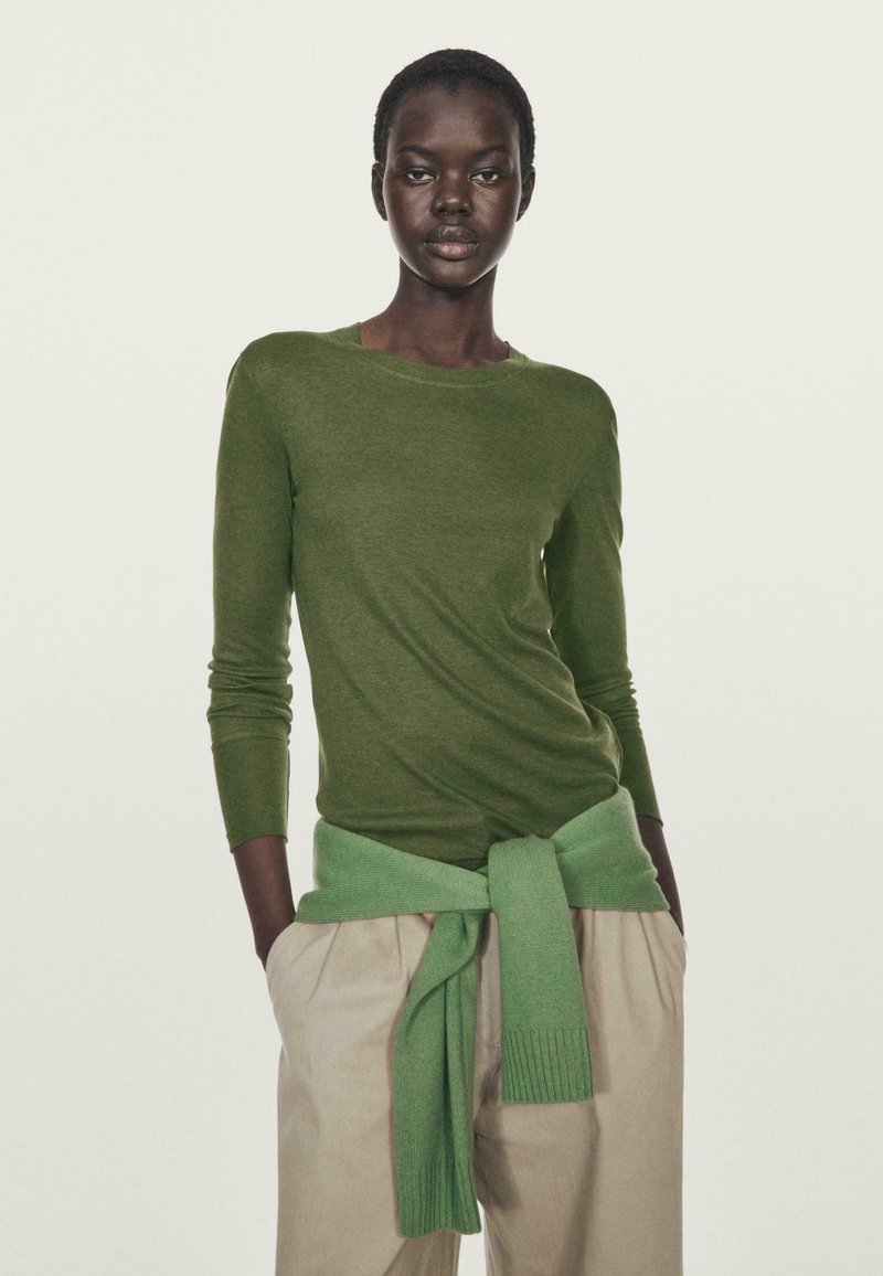Top a maniche lunghe verde oliva con scollatura rotonda, abbinato a pantaloni beige chiaro e una sciarpa lavorata a maglia verde menta annodata in vita.