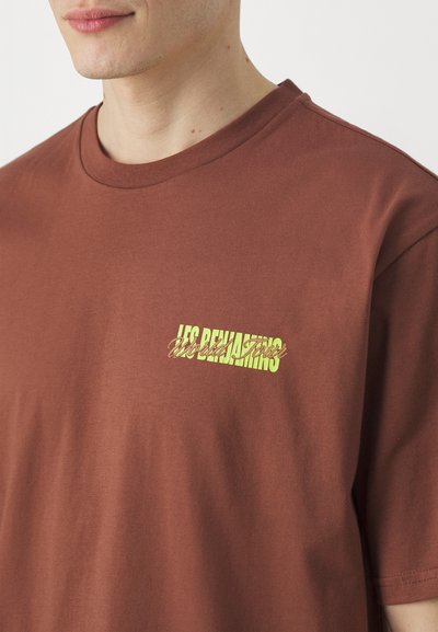 T-shirt en coton marron avec un col rond et des manches courtes. Présente un imprimé graphique jaune sur la partie gauche de la poitrine. Texture lisse.