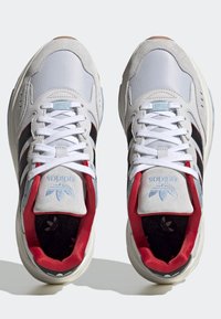 Ett par vita Adidas-sneakers med svarta ränder, röd innersula och blå logo på tungan, sedd uppifrån mot en vit bakgrund.