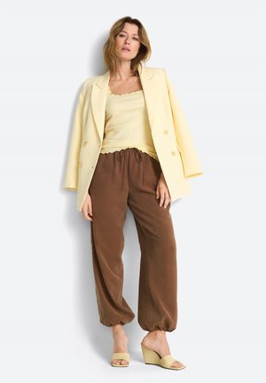 Naine, kellel on seljas helekollane jakk sobiva tanktopi ja pruuni lahtiste jalgadega retuusid elastsete pahkluude juures ning kollased kiiltaolised sandaalid, seisab valge tausta ees.