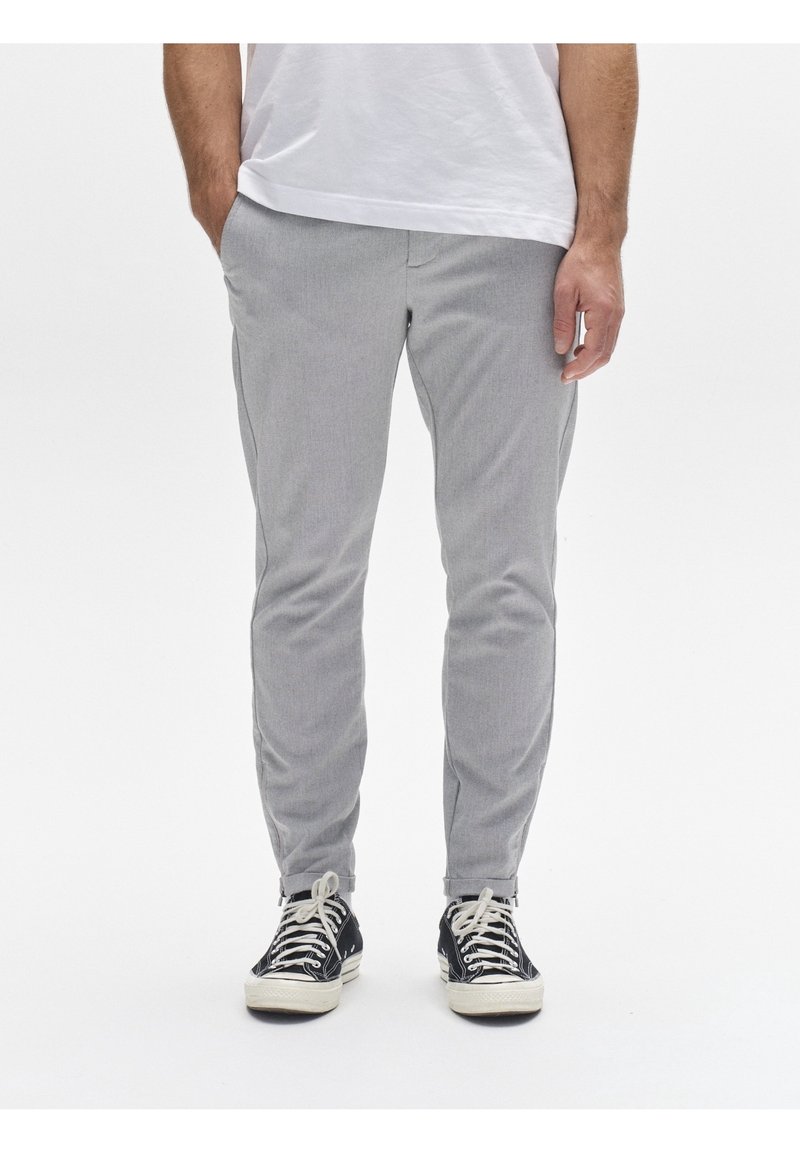 Homme portant un pantalon slim gris clair et des baskets montantes noires, debout les mains détendues le long du corps.
