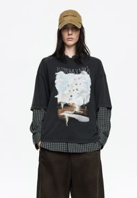 Camiseta negra de gran tamaño con una gráfica de lobos en nubes, superpuesta a una camisa de cuadros verde. Gorra oliva con el logo "Bimba y Lola". Pantalones marrones.