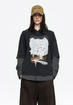 Czarny, oversize t-shirt z grafiką wilków w chmurach, nałożony na zieloną koszulę w kratę. Oliwkowy kapelusz z logo "Bimba y Lola". Brązowe spodnie.