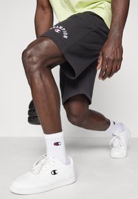 Svarta shorts med en vit "Champion"-logotyp, kombinerat med vita strumpor med logotypen och vita sneakers. Tyget verkar mjukt och sportigt.