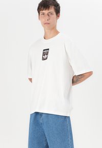 White cotton t-shirt featuring a black patch with a patterned logo centered on the front. Short sleeves and a relaxed fit. Denim shorts.