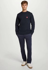 Marineblå strikke genser med rund hals og ribbestrikkede mansjetter, med en liten rød logo. Kombinert med marineblå bukser og hvite joggesko.