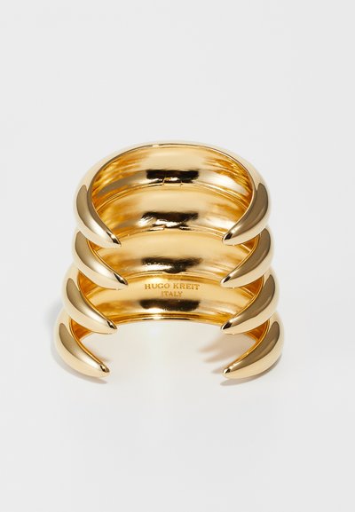 Gold-tone cuff bracelet featuring a smooth, polished finish with three curved claw-like accents. Text engraved on the inner surface.