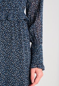 Kwiatowa sukienka w granatowym kolorze z jasnoniebieskimi i kremowymi wzorami kwiatowymi. Lekka tkanina, długie rękawy z falbaniastymi mankietami i talia podkreślona paskiem.