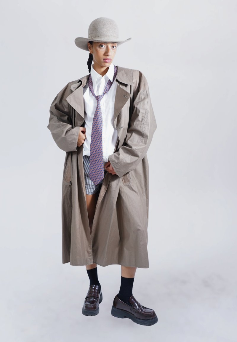 Long beige coat, white shirt, patterned tie, gray shorts, black socks, and chunky black shoes. Gray felt hat completes the look.