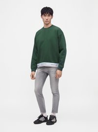 Jeune homme debout portant un sweat-shirt vert foncé, un jean skinny gris clair déchiré, des baskets noires et des chaussettes blanches sur un fond blanc uni.