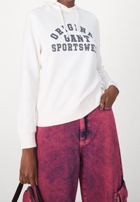 Sudadera blanca con texto en navy que dice "Original Gant Sportswear," combinada con pantalones vaqueros rosa brillante con una textura desgastada y cremalleras.