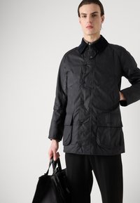 Veste imperméable noire avec des fermetures à boutons, deux poches avant et un col contrasté. Portée avec un pantalon noir et tenant un sac noir.