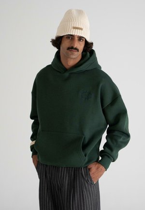 Man wearing a cream knit beanie, dark green hoodie with front pocket, and gray pinstriped trousers, standing with hands in pockets.