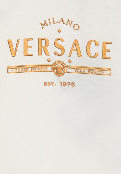 Balts kokvilnas t-krekls ar zelta metalizētu tekstu: "MILANO VERSACE" virs "NEVER FORGET YOUR ROOTS" un "EST. 1978" dekoratīvā fontā.