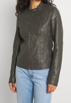 Kvinde iført mørkegrøn læderjakke med lynlås og to lynlåslommer på brystet samt lyseblå jeans, stående mod en lys baggrund.