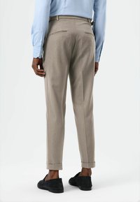 Beige Hose mit maßgeschneidertem Schnitt, ausgestattet mit zwei Gesäßtaschen, gekrempelten Säumen und einem glatten, strukturierten Stoff. Kombiniert mit schwarzen Loafers.