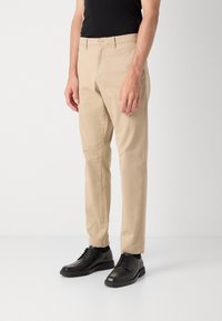 Chino beige con una texture liscia, design a gamba dritta, caratterizzati da una chiusura frontale con bottone e tasche laterali, abbinati a scarpe in pelle nere.