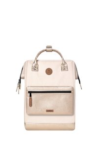 Un sac à dos couleur crème avec des accents dorés métalliques, une poche zippée texturée, un tissu lisse et deux poignées supérieures pour un transport facile.