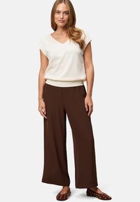 White v-neck cap-sleeve top paired with loose brown pants. Model wears brown flats with gold studs. Smooth fabric with a relaxed fit.