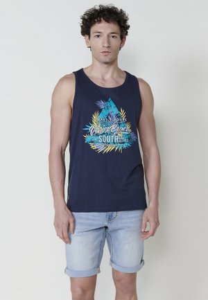 Marineblå tanktop med et grafisk design, der har tropiske blade og tekst, parret med opkrøllede lyse denimshorts. Glat bomuldsstruktur.