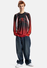 Melna un sarkana krekls ar garām piedurknēm un gradienta rakstu, ar baltām akcentiem un logotipu, kombinēts ar brīvi pieguļošām tumši ziliem džinsiem un baltām sporta kurpēm.